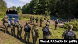 Польскія (на пярэднім плане) і беларускія (на заднім плане) памежнікі стаяць побач зь мігрантамі з Афганістану, якія сядзяць на зямлі каля невялікай вёскі Горны Уснар на польска-беларускай мяжы